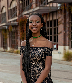 This is a photo of Alani Summers. She is a young Black woman, and in this photo she wears a black lace dress and stands in front of a brick building.