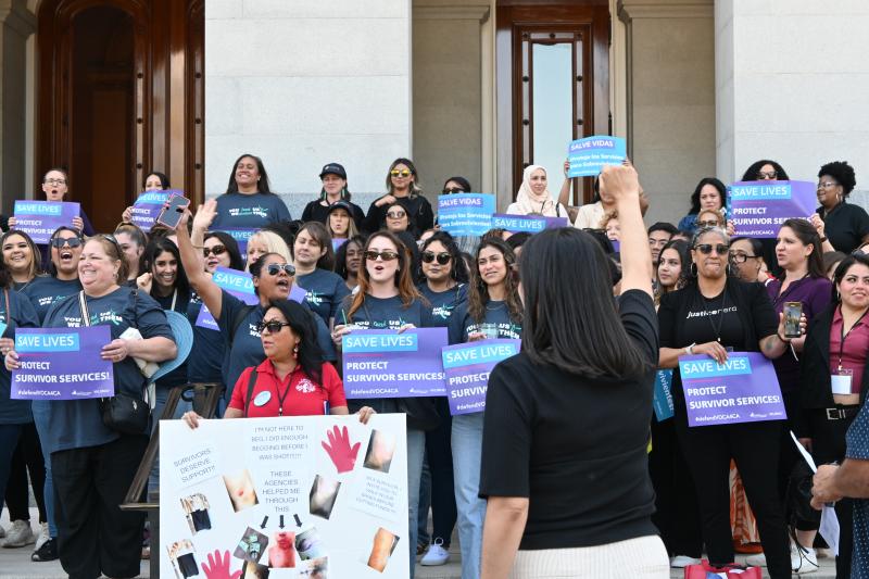 A crowd of people holding up signs, including ones that say "SAVE LIVES; PROTECT SURVIVOR SERVICES". Facing the crowd, someone raises a fist in solidarity.