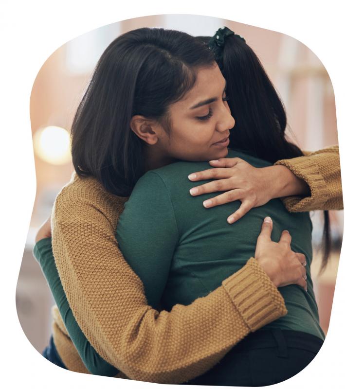 Two people are hugging. One person has shoulder-length dark brown hair and a beige sweater, and the person has long  dark brown hair and a green shirt.