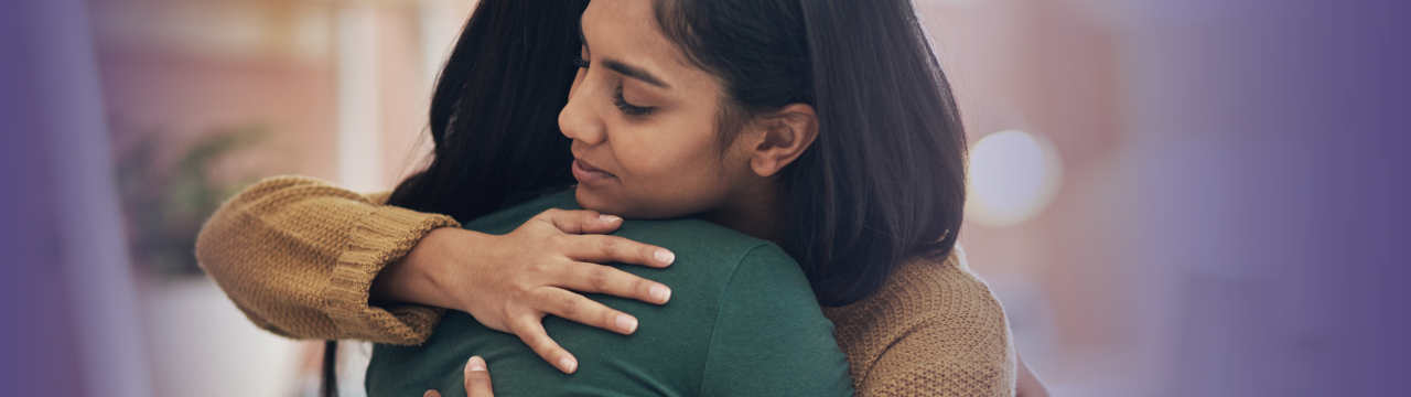 Two people are hugging. One person has shoulder-length dark brown hair and a beige sweater, and the person has long  dark brown hair and a green shirt.