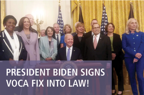 President Joe Biden sits at a desk after signing the VOCA Fix Act. Vice President Kamala Harris stands next to him, as well as a group of policymakers. In the foreground is a semi-translucent purple rectangle containing white text: "PRESIDENT BIDEN SIGNS VOCA FIX INTO LAW!"
