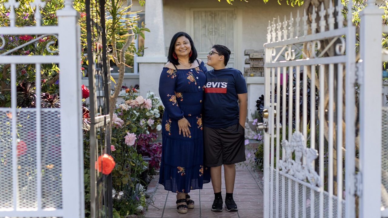 Yenni is smiling and stands in the middle of a path next to a row of flowers. Her son stands beside her and is looking up at her with a slight smile