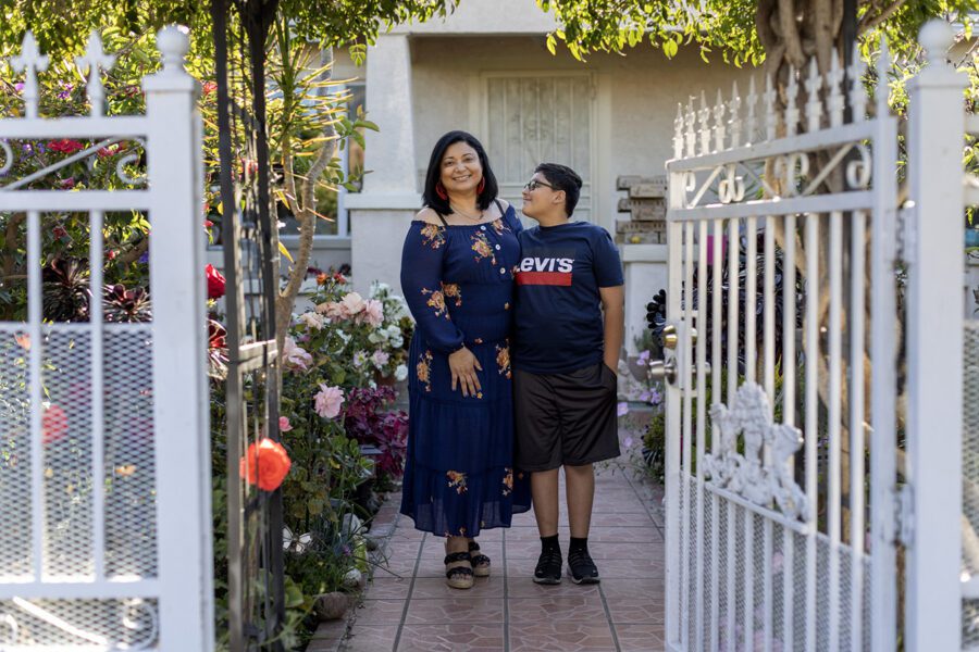 Yenni is smiling and stands in the middle of a path next to a row of flowers. Her son stands beside her and is looking up at her with a slight smile